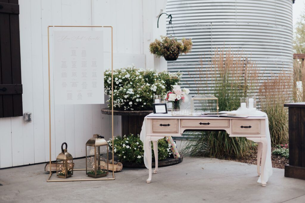 Rustic wedding decor with seating chart, lanterns, flowers, and guestbook on vintage table beside greenery backdrop.