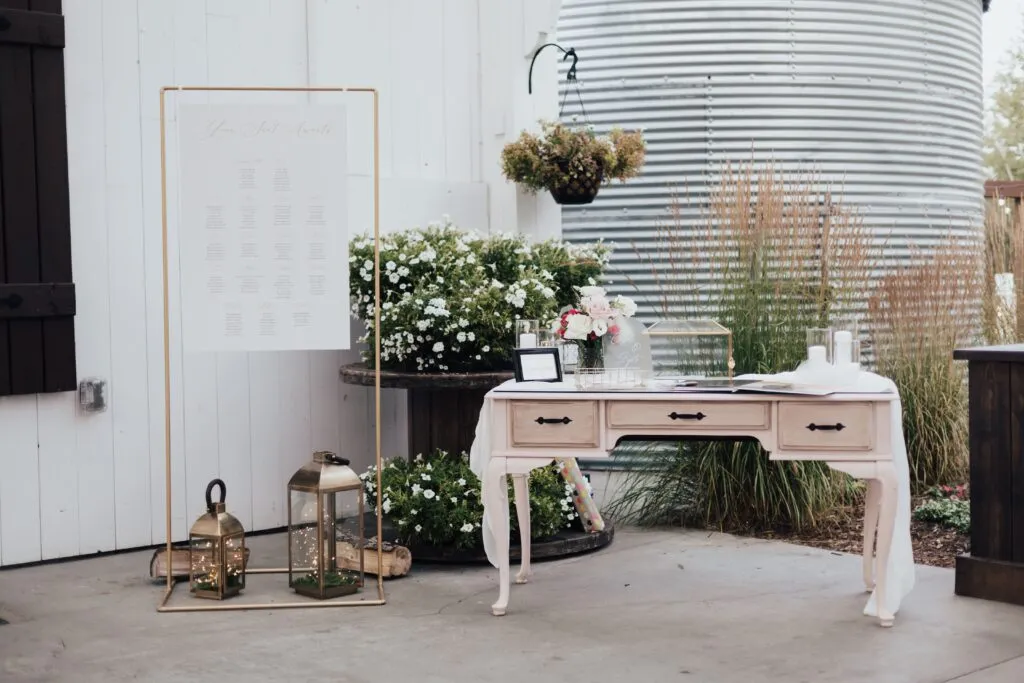 Rustic wedding decor with seating chart, lanterns, flowers, and guestbook on vintage table beside greenery backdrop.