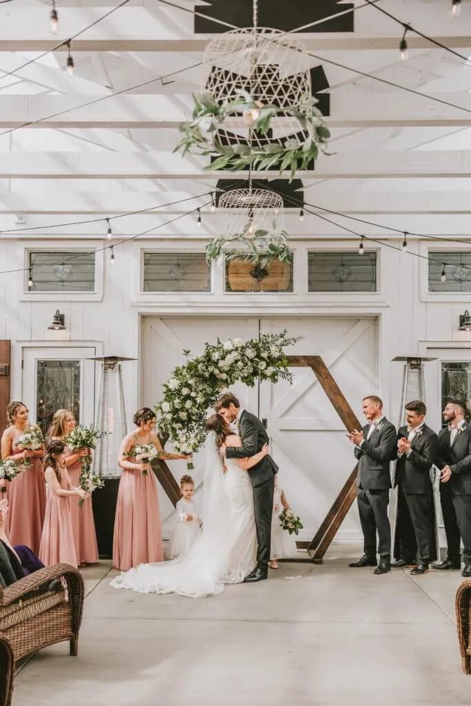 Elegant wedding ceremony with bride and groom kissing, surrounded by bridesmaids and groomsmen in a decorated venue.