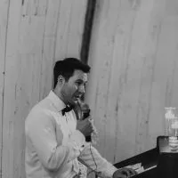 Man giving a speech at a wedding, wearing a white shirt and bow tie, speaking into a microphone.